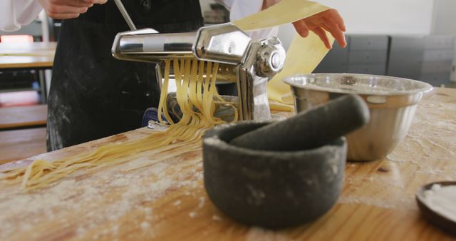 Chef rolling fresh pasta dough with a pasta machine, demonstrating culinary skills in an authentic kitchen. A small pile of flour is seen on wooden table, along with a metal bowl and a mortar and pestle, capturing the essence of traditional Italian cooking. Ideal for use in articles or blog posts about homemade pasta, culinary tutorials, Italian cuisine, or traditional cooking methods.