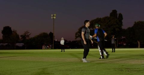 Floodlit evening cricket team bowling, running and preparing on pitch with diverse teammates