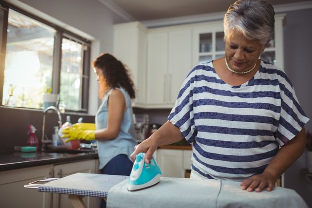 This image depicts a mother ironing clothes while her daughter washes dishes in the kitchen. It can be used to illustrate themes of family life, household chores, teamwork, and daily routines. Ideal for articles or advertisements related to home care, family dynamics, and domestic responsibilities.