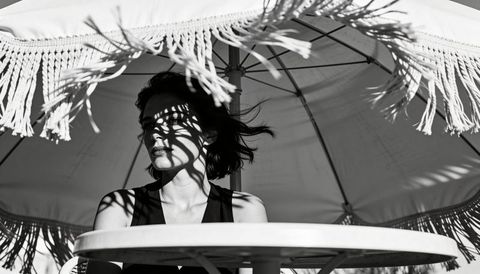 Woman sitting under fringed parasol casting dramatic shadow patterns monochrome
