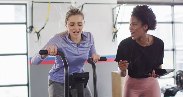 Image of diverse female fitness trainer and woman on exercise bike bumping fists at a gym. Exercise, fitness and healthy lifestyle.