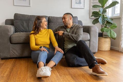 Couple sharing laughs while sitting on wood floor by gray sofa cozy home connection