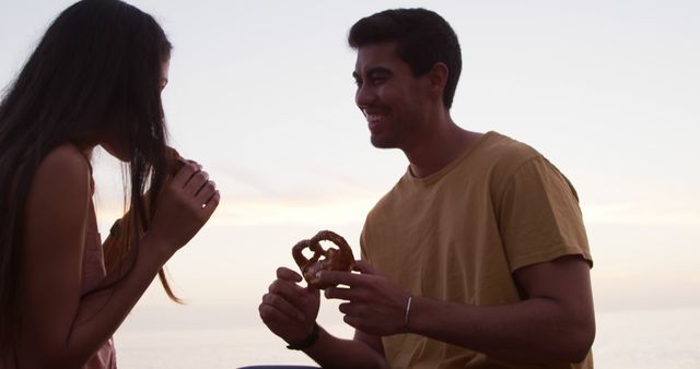 Young couple sitting by ocean during sunset, sharing pretzel and smiling. Perfect for themes around romance, leisure, happy moments, beach activities, or evening snacks.