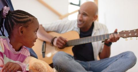 African american father playing acoustic guitar with daughter holding teddy in cozy home