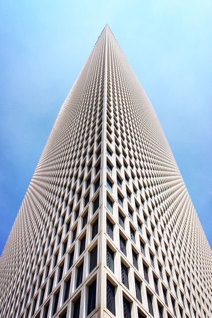 The image showcases an upward view of a modern skyscraper, accentuating its geometric design against a clear blue sky. The distinctive grid pattern of windows creates a sense of symmetry and abstract art. Ideal for use in real estate promotions, business presentations, architecture blogs, urban-themed art, or corporate websites.