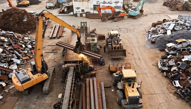 Yellow excavator cutting steel beams at busy scrap yard with heavy machinery action