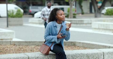 African american woman sipping coffee while checking phone on urban plaza bench in denim jacket