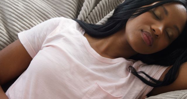 Content depicts a young Hispanic woman resting peacefully on a comfortable sofa. She is wearing a casual outfit, including a light pink t-shirt. This can be used in advertising for relaxation, home decor, lifestyle blogs, or wellness. Suitable for illustrating moments of tranquility and comfort.