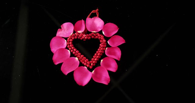 Rose petals are arranged in a heart shape, surrounded by a circle of red beads on a black background, with copy space. The composition symbolizes love and romance, often used in themes related to Valentine's Day or anniversaries.