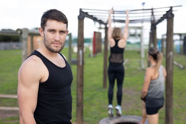 This image shows a fit man in a black vest at an outdoor gym during a bootcamp training session. Two women are seen in the background using monkey bars. Ideal for promoting fitness programs, outdoor exercise routines, and healthy lifestyle campaigns.