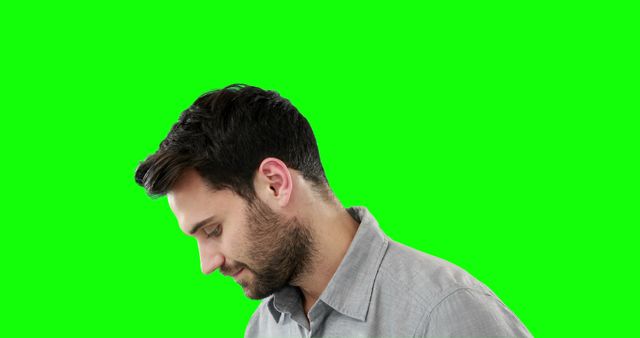 Young man with a thoughtful expression looking down against a green screen background. Uses include creating diverse multimedia content, green screen editing projects, promotions, and introspection concepts.