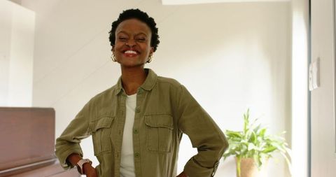 Smiling african american woman standing hands-on-hips by piano in sunlit home interior