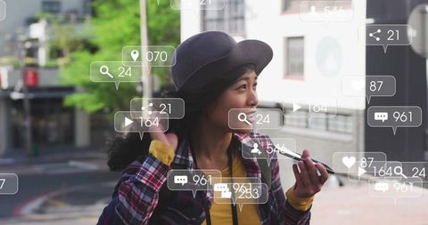 Talking asian woman holding smartphone, touching hat on city sidewalk with social overlays