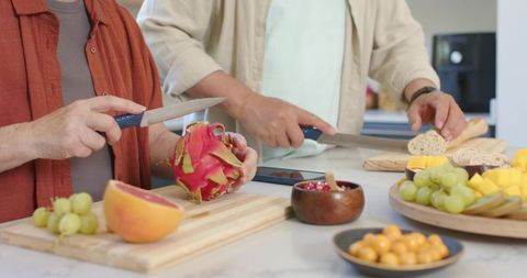 Mature asian couple slicing dragon fruit and bread on kitchen countertop using tablet