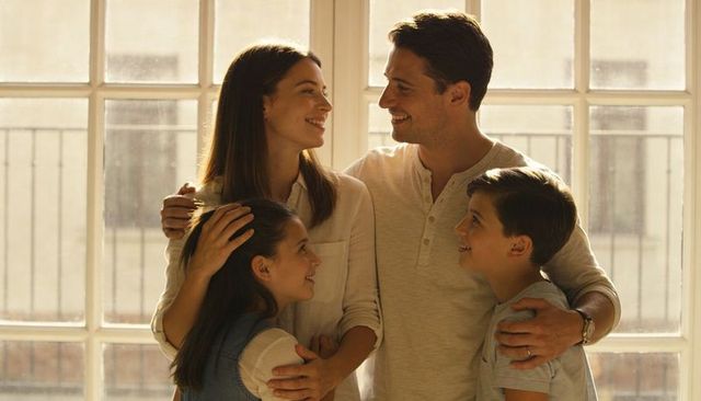 Sunlit family embracing by multi-pane window, parents smiling with daughter and son, warm