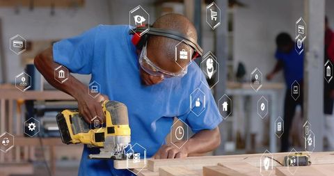 Carpenter wearing safety goggles cutting wooden plank with yellow jigsaw and digital icons