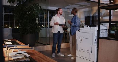 Colleagues discussing near office printer in modern workspace at night