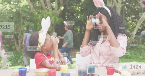 Mother and daughter painting easter eggs in garden while wearing bunny ears and laughing