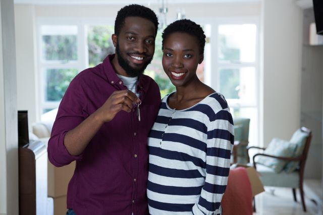 Happy young African American couple holding key to their new home and smiling. They are standing indoors, in the living room, with boxes in the background, suggesting moving-in day. Ideal for use in real estate advertisements, articles about first-time homebuyers, or lifestyle blogs focusing on homeownership and family.