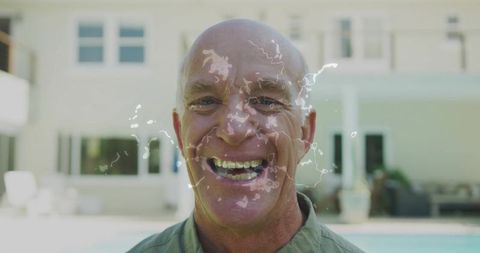 Smiling senior man splashing water at poolside backyard patio enjoying sunny summer day