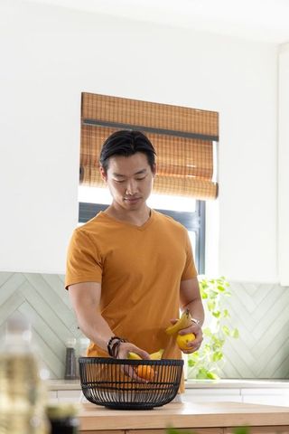 Asian man arranging bananas and oranges in wire fruit bowl on bright minimalist kitchen counter
