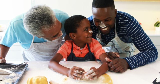 African American grandfather, father, and son enjoy baking together in a bright kitchen. They are smiling and share a special bonding moment while covered in flour. This image is perfect for promotional materials on family, bonding, cooking, and lifestyle topics. It can be used in advertisements, blogs, or social media posts focused on family values, fun activities, and cooking together.