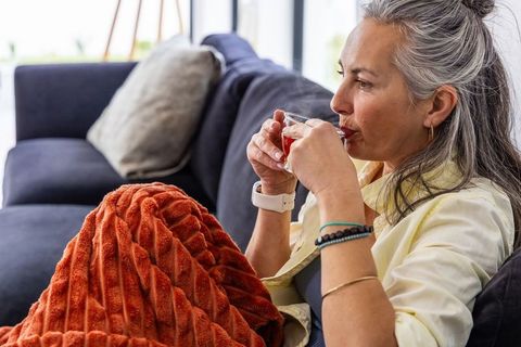 Mature woman relaxing on sofa sipping tea wrapped in textured rust blanket