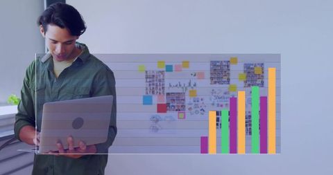 Young man holding laptop analyzing growth chart overlay in creative office workspace