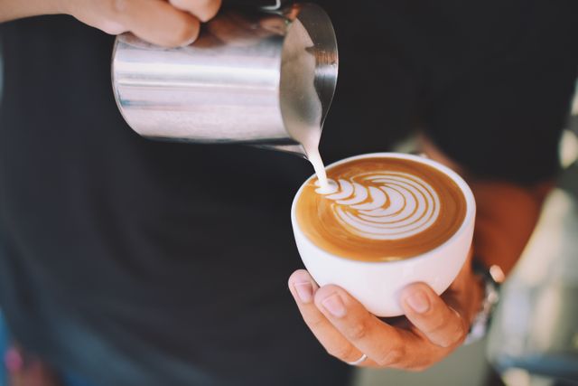 Barista pouring milk into coffee cup creating latte art in sleek swirl pattern. Ideal for websites, blogs, or social media posts related to coffee culture, cafes, barista skills, coffee recipes and drink preparations.