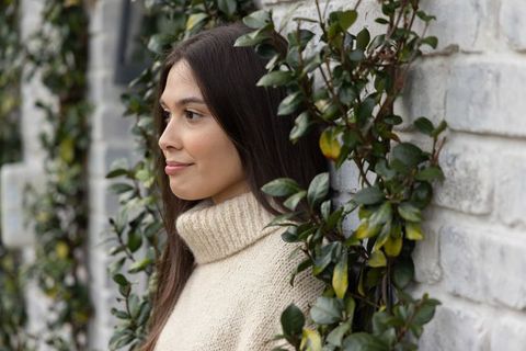 Contemplative woman in cream turtleneck sweater leaning on white brick wall amid green vines