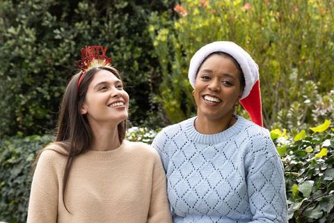 Smiling diverse friends wearing santa hat and merry headband sitting on garden bench celebrating hol