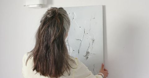 Mature woman hanging textured abstract canvas in minimalist interior near wall sconce