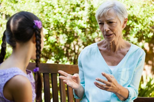 Senior woman talking with granddaughter while sitting on wooden bench at backyard