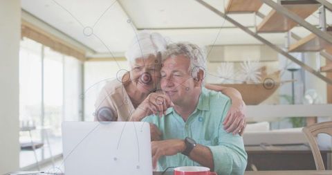 Senior couple embracing and pointing at laptop in bright open-plan living room