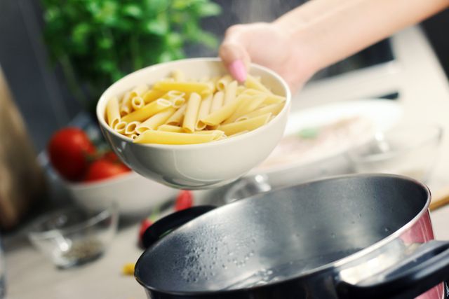 Hand holding bowl of uncooked penne pasta above boiling pot in kitchen. Background includes fresh ingredients such as tomatoes and greens. Ideal for articles on cooking, recipes, food blogs, or kitchen-related products.