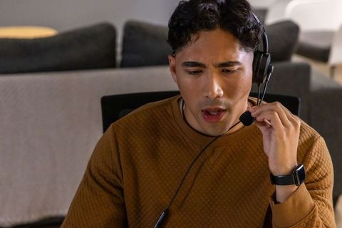 Asian man speaking into headset in cozy home office wearing brown pullover and smartwatch