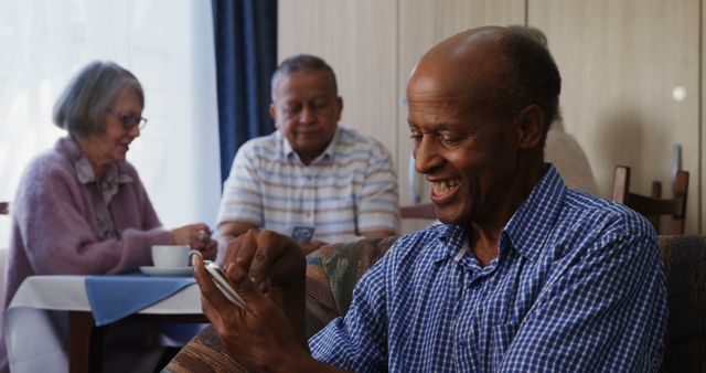 Senior man in casual clothing smiling while using smartphone, with elderly friends sitting and chatting in background. Can be used for depicting technology adoption among seniors, conveying positive emotions and leisure activities in retirement communities, or illustrating modern tech engagement across age groups.