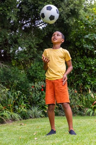 African american boy juggling soccer ball on garden lawn in yellow tee and orange shorts