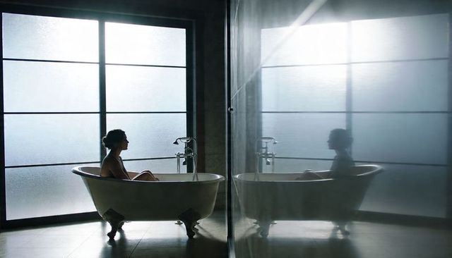 Woman sitting in clawfoot tub soaking by frosted window with mirrored partition silhouette