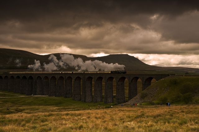 Historic steam train crossing over a grand viaduct during sunset. Dramatic sky with rolling hills in the background. Ideal for travel, adventure, history, and transportation themes. Captivates viewers with its blend of natural beauty and human engineering.