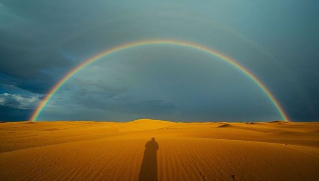 Vivid double rainbow arcing over golden desert dunes casting long human shadow