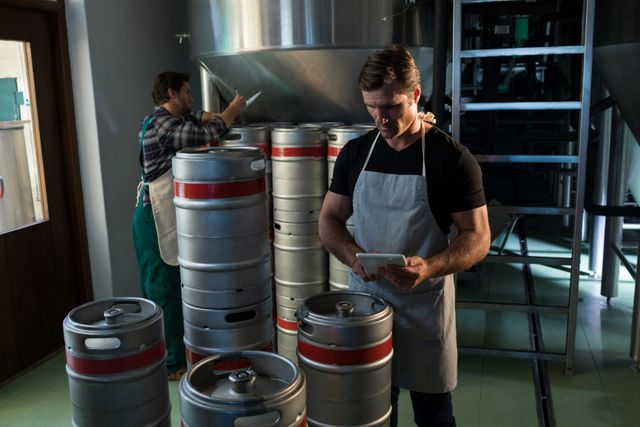 Workers in a brewery warehouse managing inventory of kegs. One worker is using a tablet while another is checking kegs. Ideal for illustrating industrial work environments, logistics, teamwork, and inventory management.
