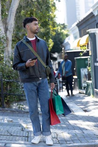 Indian man shopping and checking smartphone on sunlit urban sidewalk with colorful bags
