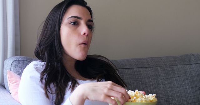 This image shows a young woman sitting on a couch while eating popcorn, focusing on casual relaxation and leisure activities at home. The setting reflects a typical living room, hinting at cozy, everyday life. This image is suitable for promoting home entertainment, relaxation products, or lifestyle blogs.