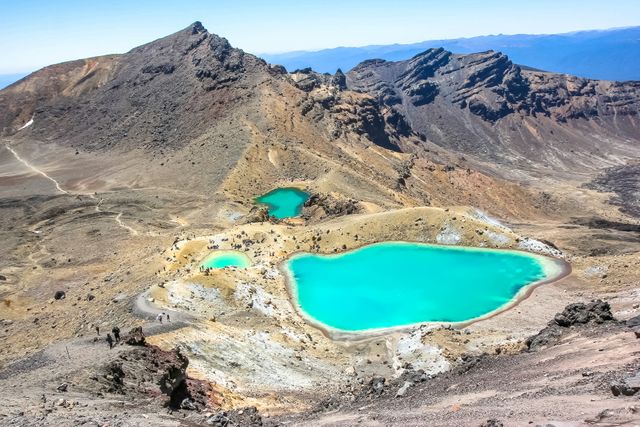 The image features the stunning Emerald Lakes situated in Tongariro National Park, New Zealand. These lakes are characterized by their vibrant turquoise colors, surrounded by a rugged, volcanic landscape. You can spot hikers making their way across the terrain, emphasizing the adventurous spirit of the park. This image can be used for travel promotions, adventure tourism marketing, environmental conservation campaigns, and outdoor enthusiast blogs.
