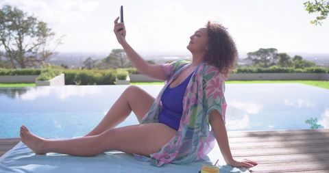 African american woman relaxing by sunlit infinity pool taking selfie in floral cover-up