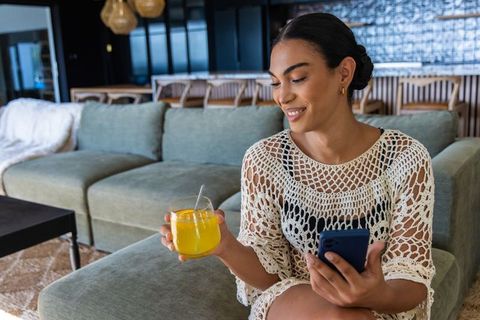 African woman relaxing on sofa holding orange juice and smartphone in modern living room