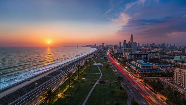 Coastal highway curving past palm-lined park at sunset with city skyline and light trails