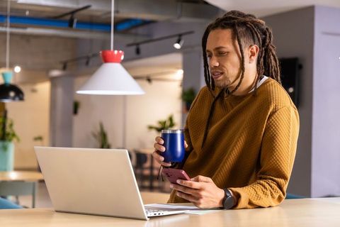 African American man leaning on coworking table checking phone holding tumbler with laptop