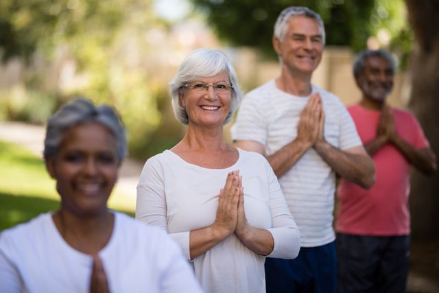 Senior individuals standing in a park, meditating with hands in prayer position. They are smiling and appear relaxed, enjoying the outdoor setting. This image can be used for promoting wellness, fitness, and healthy lifestyle among elderly people. It is also suitable for advertisements related to meditation, mindfulness, and community activities for seniors.